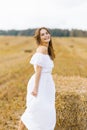 A fair-haired young woman in a white dress in a field with stacks of straw enjoys a summer day and smiles Royalty Free Stock Photo