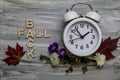 FALL BACK Daylight Saving Time concept with white alarm clock and fresh fall flowers foliage on hand painted gray wood board flat Royalty Free Stock Photo