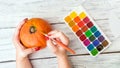 Fall Halloween crafts. Female hands with orange decorative pumpkin and bright paint with brush on white wooden background. Jack O