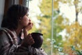 Fall melancholy mood concept. Wistful woman sitting near window and looking at autumn landscape outside in rainy day. Copy space