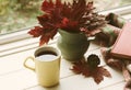 Fall Still Life Arrangement with Red Maple Leaves in a Vase, Yellow Coffee Mug, and a Book on White Board Table in front of a Wind
