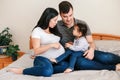 Family Asian Chinese pregnant woman and Caucasian man with toddler girl sitting on bed at home. Mother and father talking