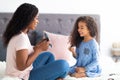 Family beauty day. Cute little black girl applying lipstick while her mom holding mirror on bed at home