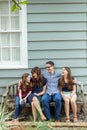 A family of four with a mother and father and two daughters sitting on a bench outside a blue cottage house