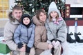 A family of four poses in winter in the courtyard of the house against the backdrop of a Christmas tree and a large
