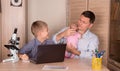 Family lifestyle concept. Smiling boy doing homework using laptop. Father with a newborn baby in his hands helping his laughing s Royalty Free Stock Photo