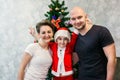 Family mom, dad and son in Santa costume are standing near the Christmas tree in their apartment and looking into the camera Royalty Free Stock Photo