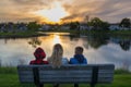 Family, mom with her two kids from back watching the sunset, sitting on a bench near a pond Royalty Free Stock Photo