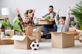 Family: playful siblings sitting in carton boxes with raised arms and enjoying relocation in new flat on background of parents