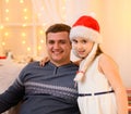 Family portrait - father and daughter in home interior decorated with holiday lights and gifts, dressed in santa hat for new year