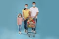 Family with protective masks and shopping cart full of groceries on light blue background Royalty Free Stock Photo