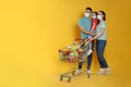 Family with protective masks and shopping cart full of groceries on yellow background. Space for text Royalty Free Stock Photo