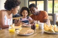 Family Shot With Parents And Daughter At Home Having Breakfast Spreading Jam On Bread At Table