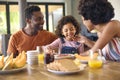 Family Shot With Parents And Daughter At Home Having Breakfast Spreading Jam On Bread At Table