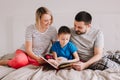 Family of three people sitting on bed in bedroom reading book. Mother, father and boy son at home spending time together. Parents Royalty Free Stock Photo