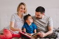 Family of three people sitting on bed in bedroom reading book. Mother, father and boy son at home spending time together. Parents Royalty Free Stock Photo