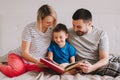 Family of three sitting on bed in bedroom reading book. Mother, father and a boy son at home spending time together. Parents Royalty Free Stock Photo