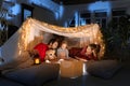 Family time. Mother, father and daughter lying inside self-made hut, tent in room in the evening and reading Royalty Free Stock Photo