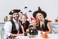 family in various halloween costumes at table with black pot with treats in kitchen