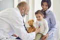 Family with a visit to the doctor in the clinic office. Senior doctor with a stethoscope listens to a little girl with