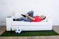 Fanatic football fan lying on couch sofa with ball on green grass carpet emulating soccer stadium pitch mocking player in pain