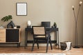 Fancy interior design of home office space with stylish chair, desk, commode, black mock up poster frame, laptop, book, desk.