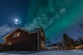 Fantastic winter landscape with wooden house with light in window in snowy mountains and Northern light in night sky. Christmas Royalty Free Stock Photo