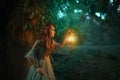 Fantasy elf princess woman walks in dark night forest, holding glowing lantern in hands. Redhead fairy girl. Summer Royalty Free Stock Photo