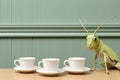 A fantasy grasshopper sitting on top of a table next to three coffee cups. AI.