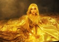 fantasy woman blonde in long golden evening shine glow dress lies in satin fabric on floor, beauty face looks at camera Royalty Free Stock Photo