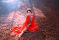 Fantasy young woman in red oriental silk dress sits on fallen orange leaves. Lady Princess in gold crown, rubies. Queen