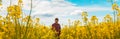 Farm worker wearing red plaid shirt and trucker`s hat standing in cultivated rapeseed field in bloom and looking over crops