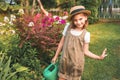 Farmer girl in summer straw hat. Little gardener farming, watering flowerbed with pink flowers, having fun in garden. Big green