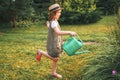 Farmer girl in summer straw hat. Little gardener farming, watering flowerbed with pink flowers, having fun in garden. Big green