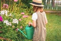 Farmer girl in summer straw hat. Little gardener farming, watering flowerbed with pink flowers, having fun in garden. Big green Royalty Free Stock Photo