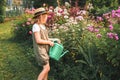 Farmer girl in summer straw hat. Little gardener farming, watering flowerbed with pink flowers, having fun in garden. Big green