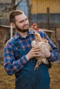 Farmer handsome smiling european caucasian rural portrait in countryside with beard, shirt and overalls looks at chicken with