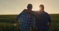 The farmer hugs his adult son and looks forward together to the wheat field where the sun is setting. Family business