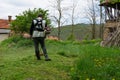 A farmer is mowing tall grass with a manual grass cutter behind his rural house on a spring day. The man is wearing boots Royalty Free Stock Photo