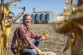 Farmer in Cornfield With Freshly Harvested Corn Cob Against Tractor and Grain Silo