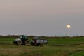 Farmer with tractor and trailer cultivating fields under a full moon at sunset