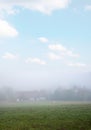 Farmstead in foggy landscape, grazing horse on the meadow, light blue sky with clouds Royalty Free Stock Photo