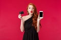 Fascinated and excited good-looking european woman with long curly ginger hair, wear black dress, holding mobile phone Royalty Free Stock Photo