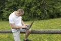 Fascinated man sitting on a wooden fence and works behind the laptop near the fields and pine forests