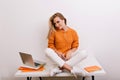 Fascinating fair-haired girl in sport shoes sitting on desk and looking with playfully smile after work with laptop