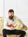 Fascinating man dressed in refined manner and reading book on upholstered sofa in bright and luxurious living room. Man