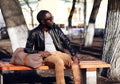 Fashion african man wearing a sunglasses and black leather jacket sits on a bench in the park Royalty Free Stock Photo