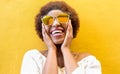 Fashion african woman smiling and wearing sunglasses with yellow ochre background - Black afro girl having fun - Focus on face - Royalty Free Stock Photo