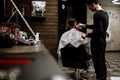 The fashion barber dressed in black clothes makes a razor cut hair for a stylish black-haired man in a stylish