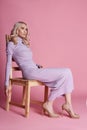Fashion Beautiful blonde woman in knitted closed long dress sitting on a chair on a pink background. Perfect hair and makeup Royalty Free Stock Photo